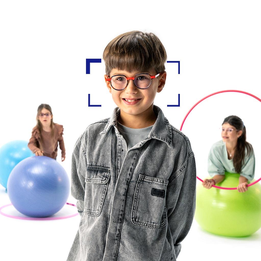 Niño de cabello castaño con gafas redondas de lentes MyoCare de ZEISS se encuentra en primer plano y sonríe a la cámara. Al fondo, dos niñas con lentes MyoCare de ZEISS jugando con aros y pelotas de gimnasia.