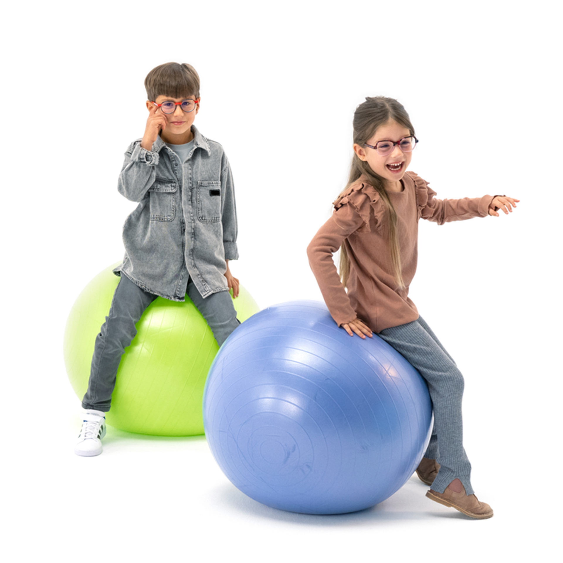 Un niño y una niña, ambos con gafas, rebotan divertidamente sobre pelotas de gimnasia.