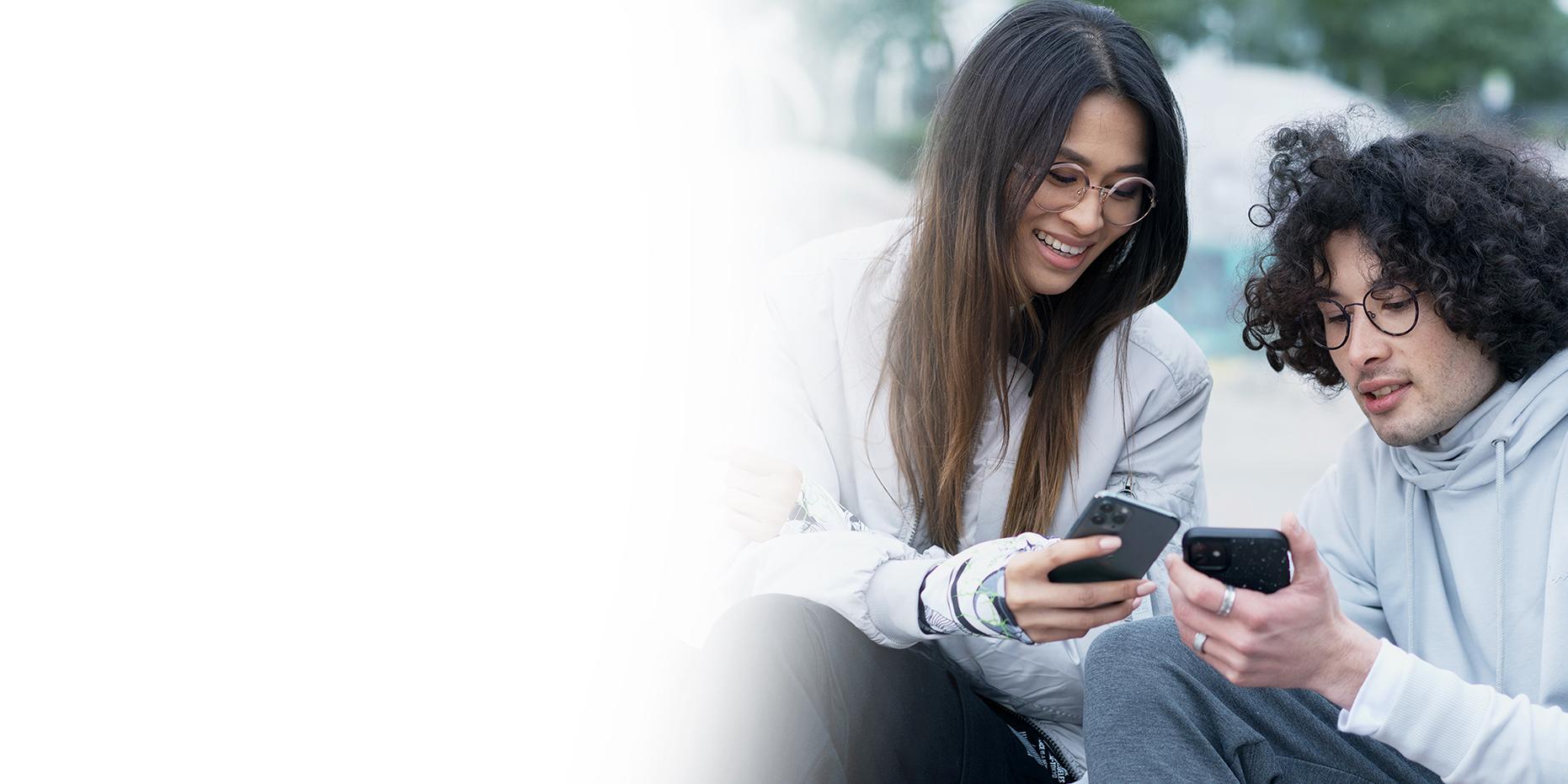 Dos jóvenes, una joven con cabello largo castaño sonriendo y un joven de cabello negro rizado con unas gafas ZEISS SmartLife, consultan sus teléfonos. 