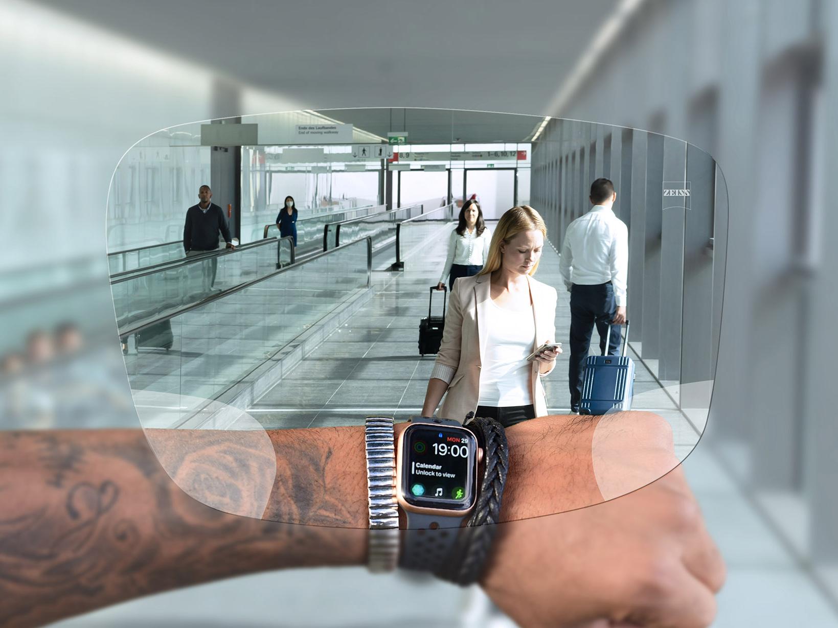 Una persona consulta su reloj a través de los lentes ZEISS Digital SmartLife. Escena típica de un aeropuerto con personas caminando en cintas mecánicas empujando sus maletas. 