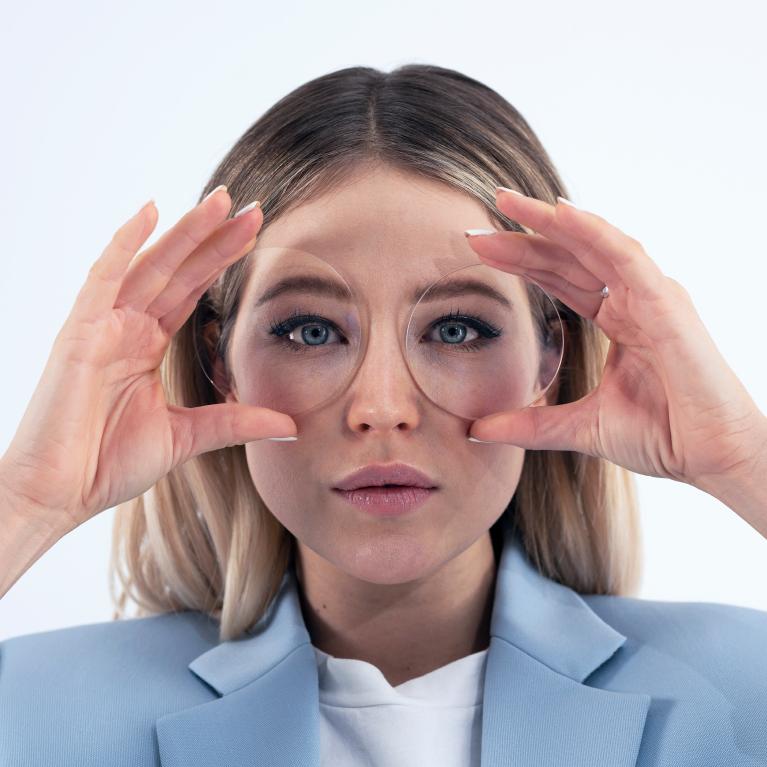 Una joven rubia sostiene unos lentes delante de los ojos para mostrar un buen aspecto sin ningún efecto raro gracias a los lentes monofocales ZEISS ClearView.