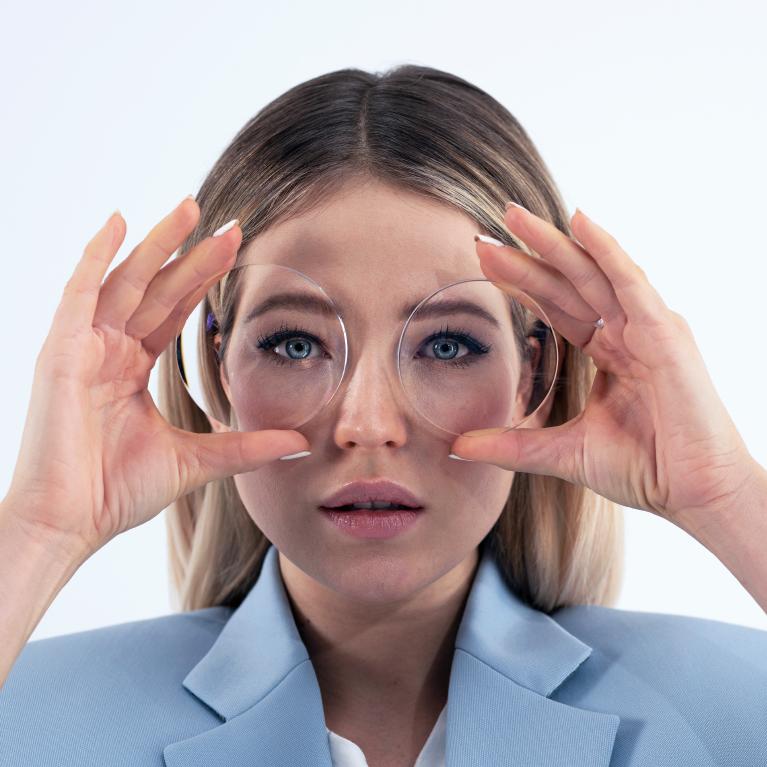 Una joven rubia sostiene unos lentes delante de los ojos para mostrar el efecto de miniojos causado por unos lentes gruesos.