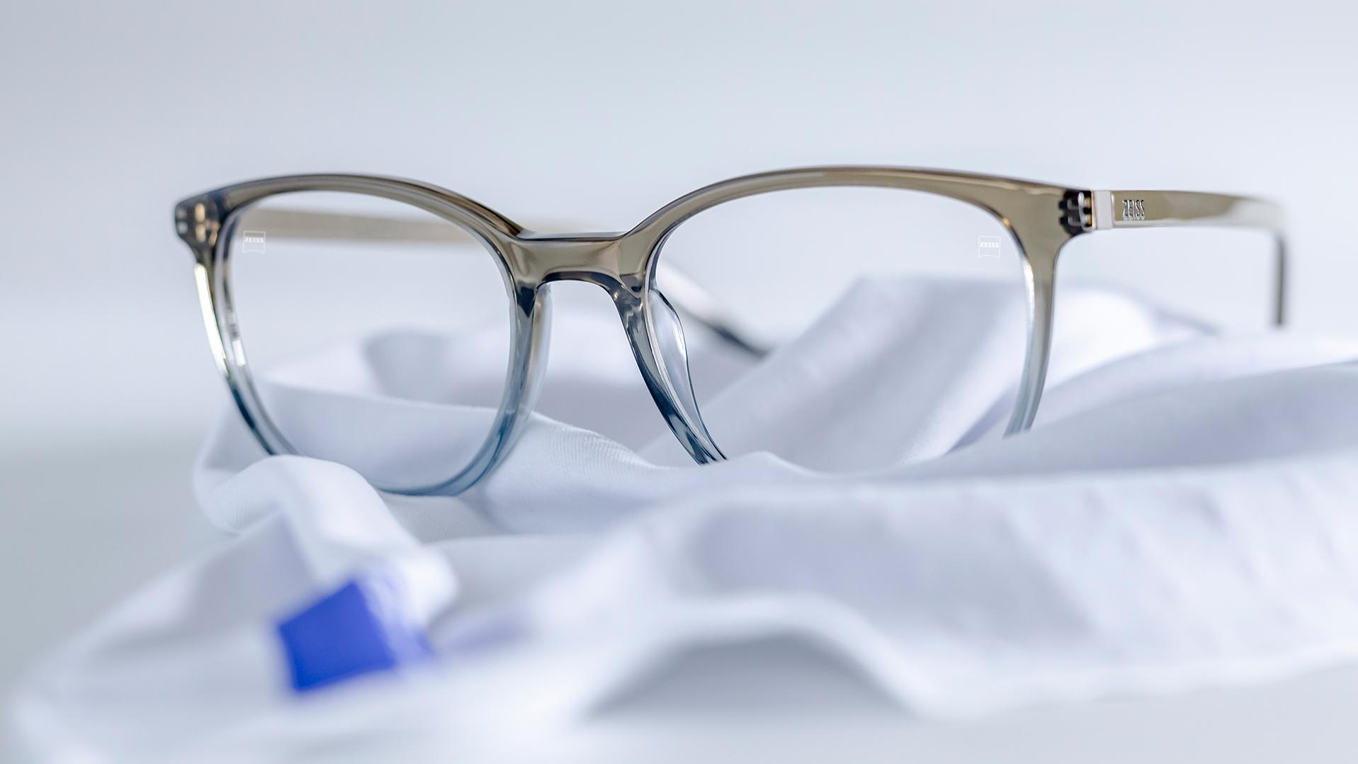 Un par de lentes con montura gris-azul y lentes ZEISS con recubrimiento DuraVision® reposan sobre un paño de microfibra blanco.
