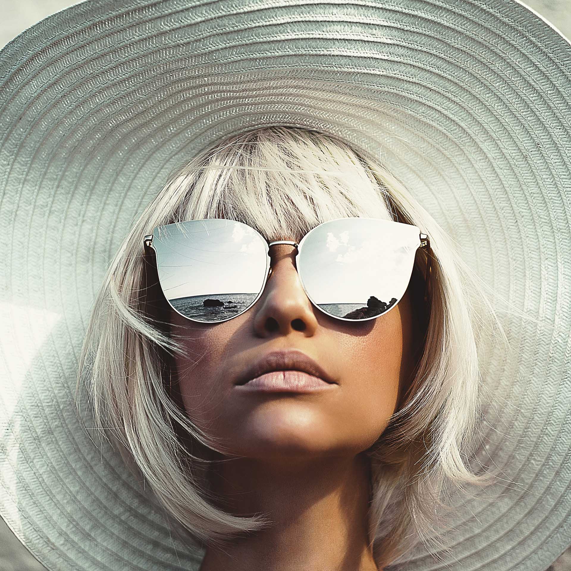 Mujer joven mira hacia arriba con enorme sombrero para el sol y grandes lentes de sol con efecto espejo.
