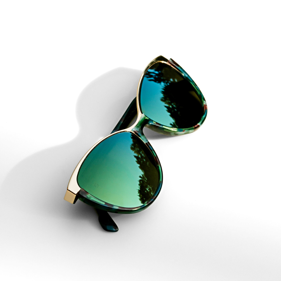 Lentes de sol tipo ojo de gato con efecto espejado en verde sobre una superficie blanca y luz incidiendo desde el lateral.