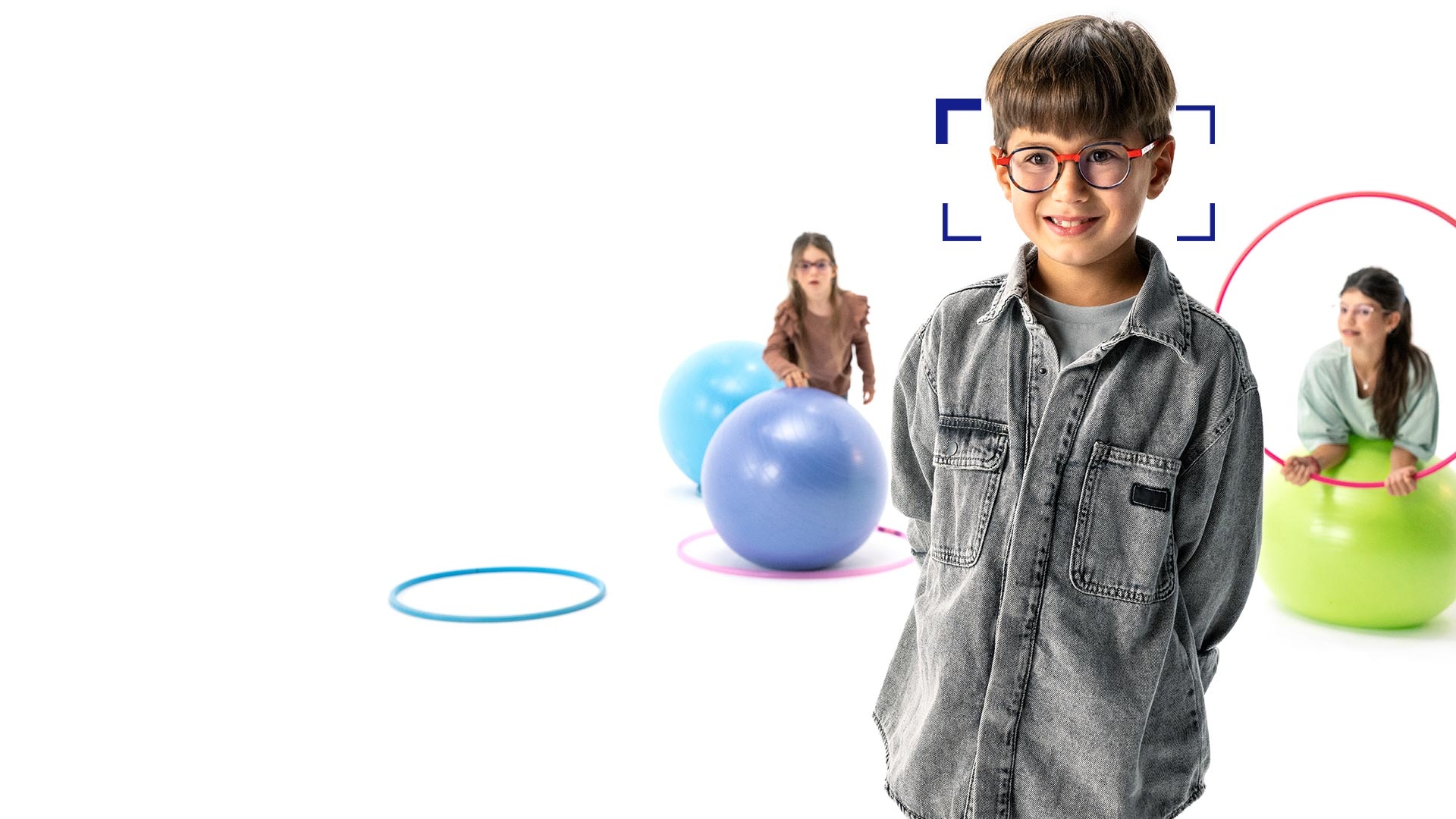 Niño de cabello castaño con gafas redondas de lentes MyoCare de ZEISS se encuentra en primer plano y sonríe a la cámara. Al fondo, dos niñas con lentes MyoCare de ZEISS jugando con aros y pelotas de gimnasia.